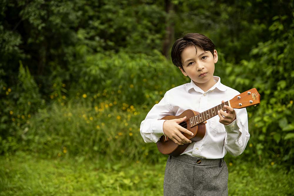 A young boy stands in front of an evergreen tree and plays his ukulele for a milestone portrait with Simply Shelayna Family photography in Syracuse NY. 