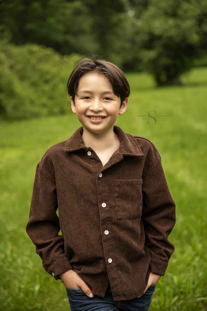 A young boys poses in an open field hedged by tall green trees with Simply Shelayna Family Photography in Oswego NY.