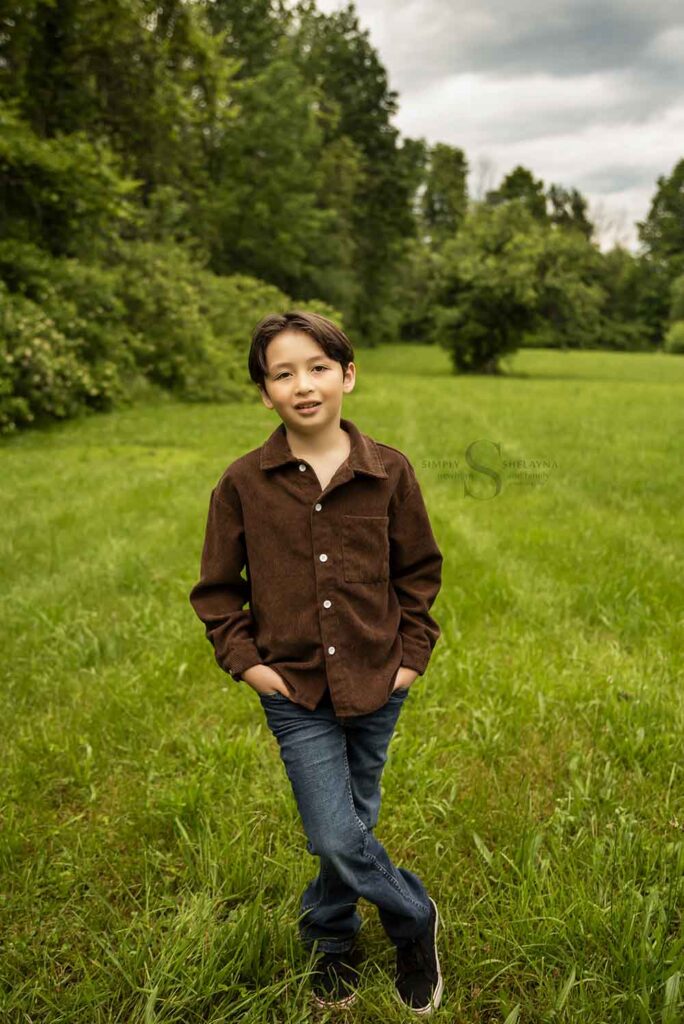 A young boys poses with his hands in his pockets and feet crossed in an open field hedged by tall green trees with Simply Shelayna Family Photography in Oswego NY.