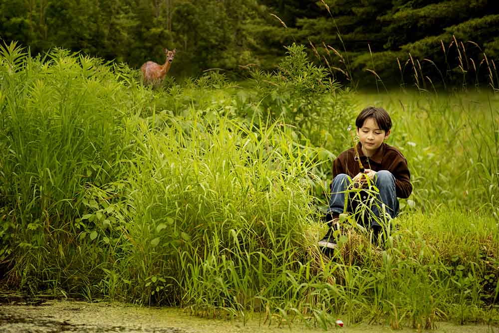 A 10 year old boy fishes by a small pond with a fawn looking on from a  distance with Simply Shelayna Child photography in Oswego NY.