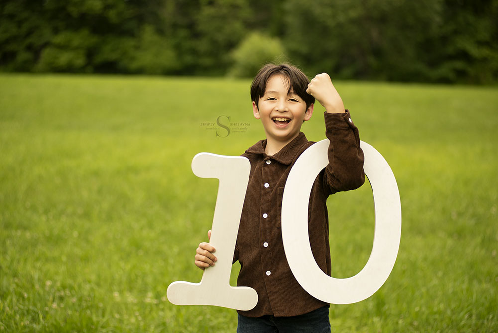 A young boy impersonates a muscular being as he holds up two digits for the number '10' with Simply Shelayna Family Photography in CNY.