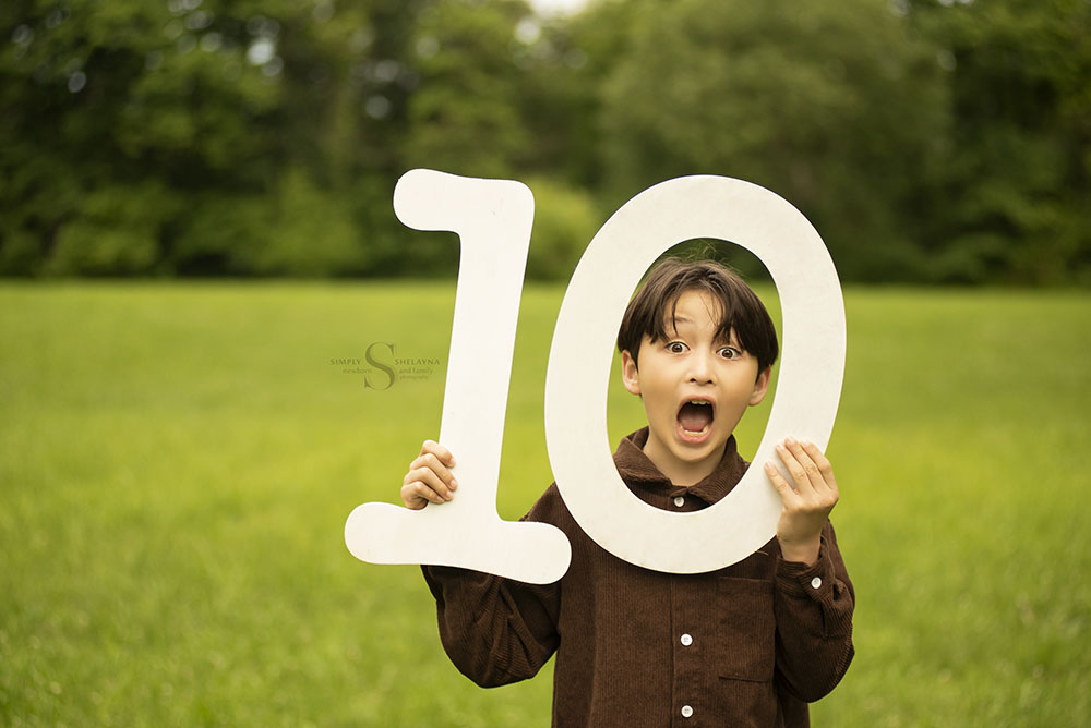 A young boy holds up a wooden 1 and 0 to signify his new age, as he make a face through the frame of the zero with Simply Shelayna Family Photography in CNY.