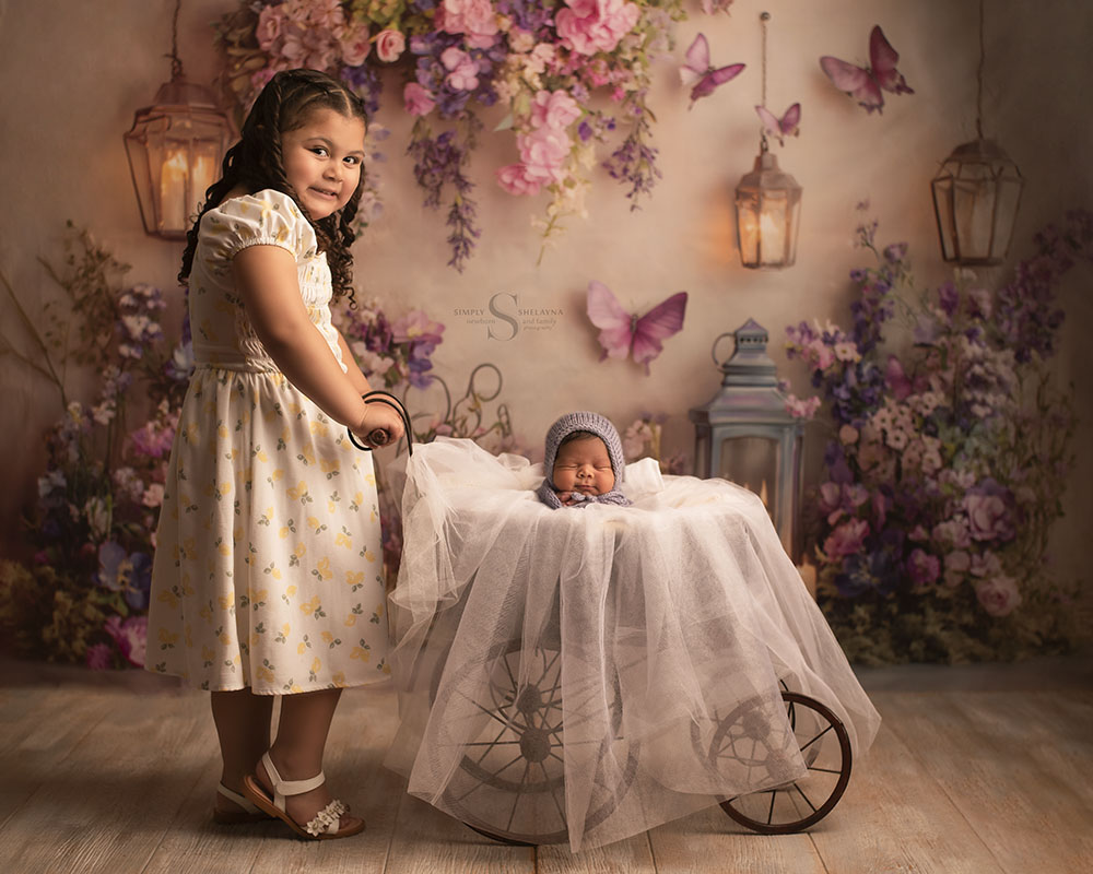 Big sister pushes baby sister in an antique carriage through a purple butterfly garden with Simply Shelayna Newborn Photography in CNY.