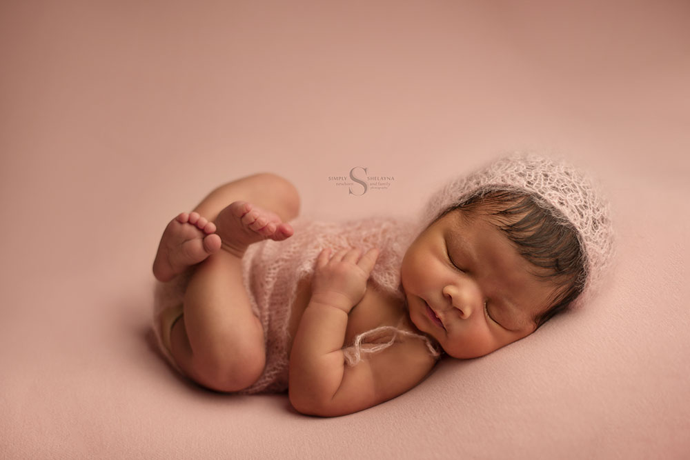 A newborn girl lies on a pink blanket and is dressed in a hand knitted pink lace onsie and bonnet for a portrait with Simply Shelayna Newborn Photography in CNY.