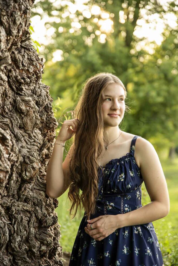 A young lady fingers her hair as she poses against an ancient weeping willow at Fall Brook in Oswego NY with Simply Shelayna Family Photography in Syracuse NY.