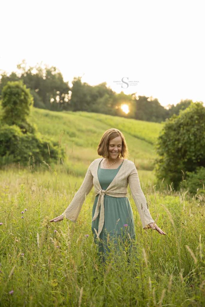 A lady dressed a sage green gown fingers her way through tall grass with the sun setting behind her with Simply Shelayna Family Photographer in Oswego NY.