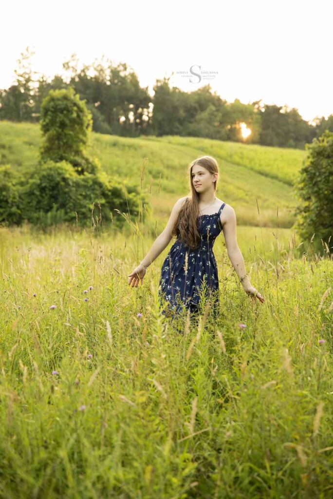 A lady dressed a blue flowered gown fingers her way through tall grass with the sun setting behind her with Simply Shelayna Family Photographer in Oswego NY.