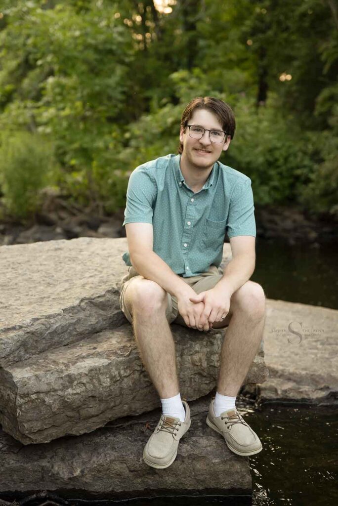 A young man sits on large rocks over Fall Brook in Oswego NY with Simply Shelayna Family photographer.