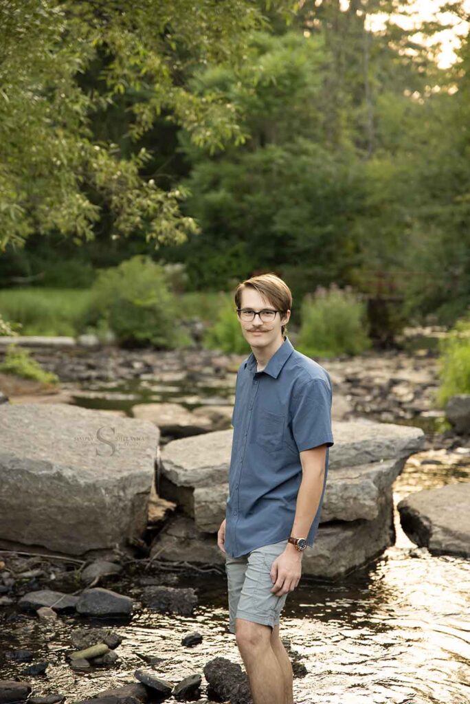 A young man walks through a shallow stony brook for a portrait with Simply Shelayna Family Photography in Oswego NY.