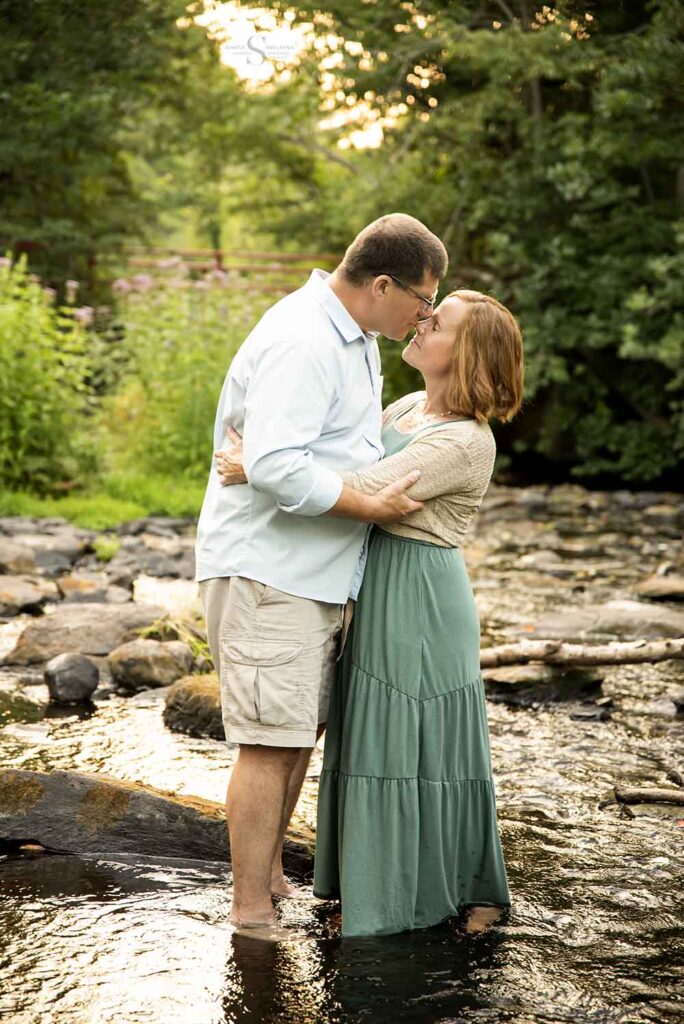 A husband tenderly kisses his wife while they stand in a stony shallow brook at SUNY Oswego's Fall Brook property with Simply Shelayna Family Photography in CNY.