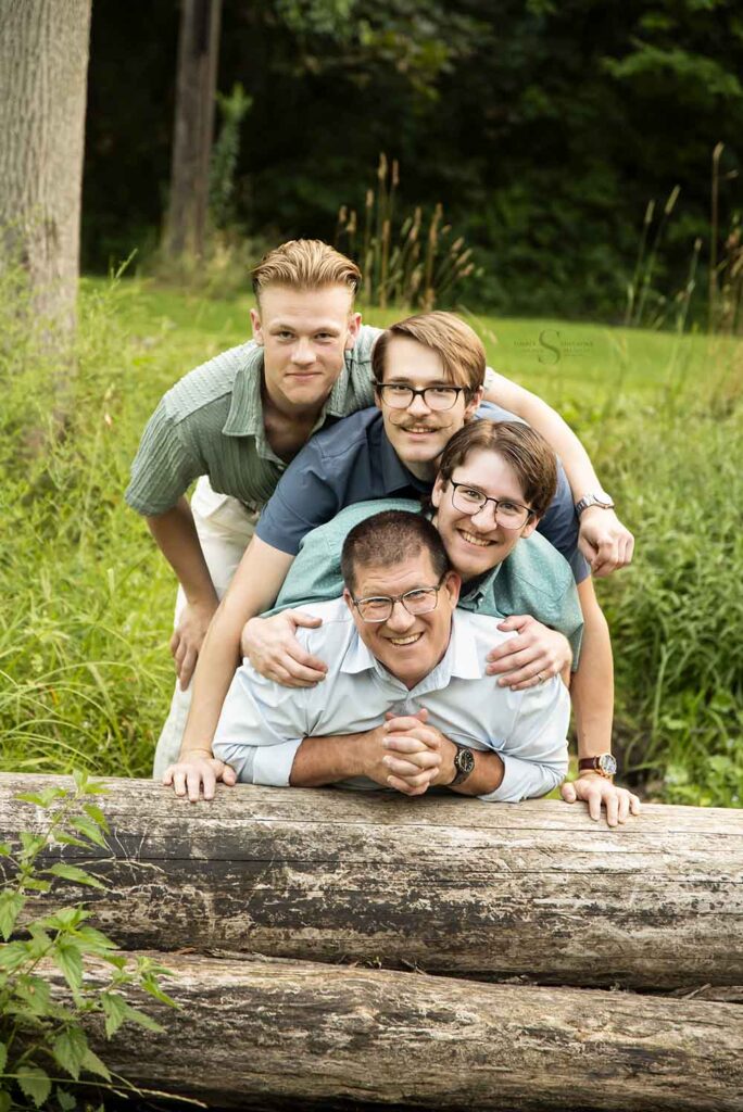 A father lays on a down log and his 3 grown sons lean into him for a portrait with Simply Shelayna Family photography in CNY.