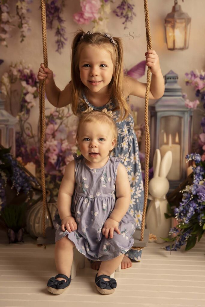 Big and little sis pose on a swing in studio for a portrait with Simply Shelayna Family Photography in Syracuse NY.