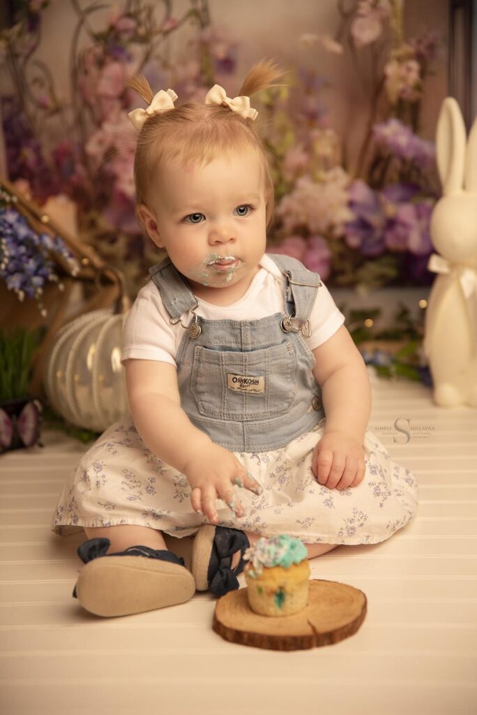 A baby girl meticulously eats her cake as she is photographed for her first birthday with Simply Shelayna Family Photography in CNY.