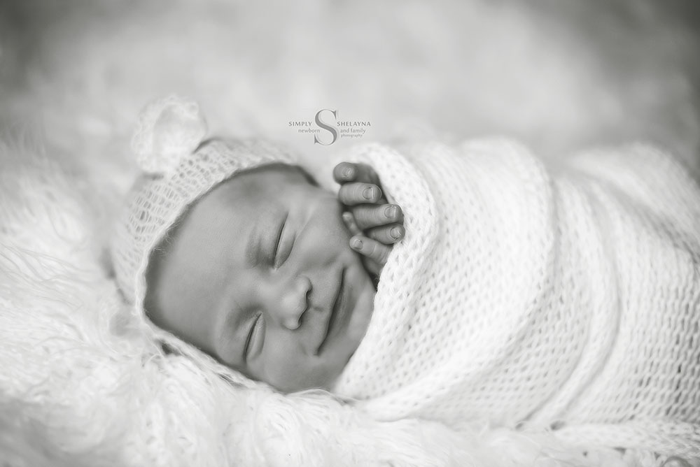 A black and white portrait of a newborn boy smiling as he is swaddled in a hand knitted wrap and bear bonnet with Simply Shelayna Newborn photography in CNY.
