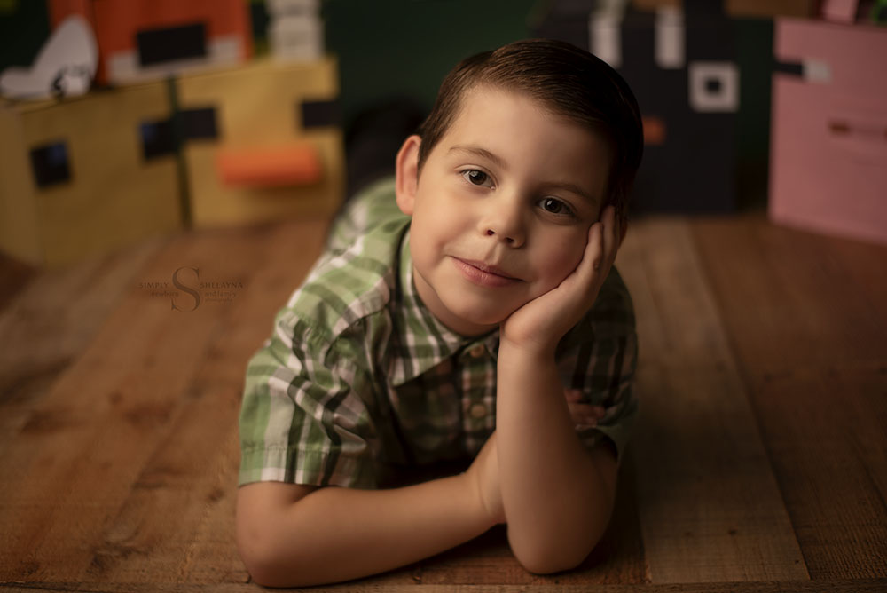 A five year old boy lies on his stomach using his hand to lean on in a Minecraft setting with Simply Shelayna Child Photography in CNY.