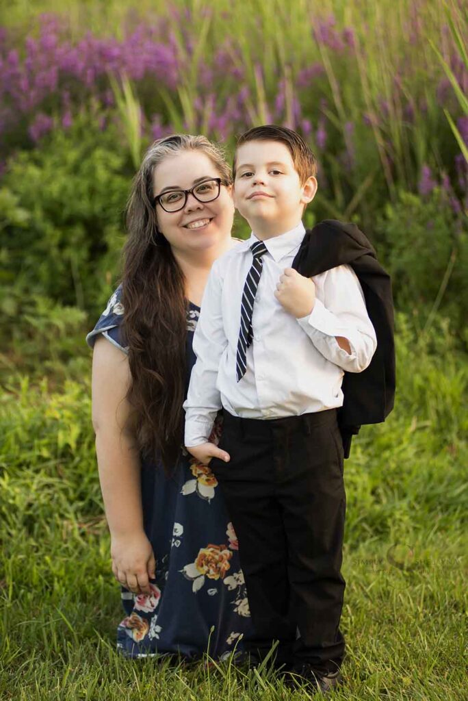 A young mother poses with her 5 year old son who is dressed in a black suit standing in front of purple wild flowers with with Simply Shelayna Family photography in Syracuse NY.